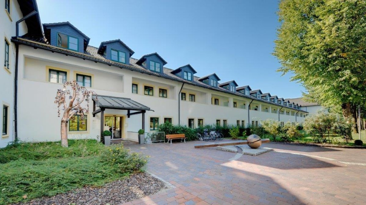Außenansicht des Asklepios Gesundheitszentrums Bad Tölz mit hellem Gebäude, begrüntem Eingangsbereich und gepflastertem Vorplatz an einem sonnigen Tag.