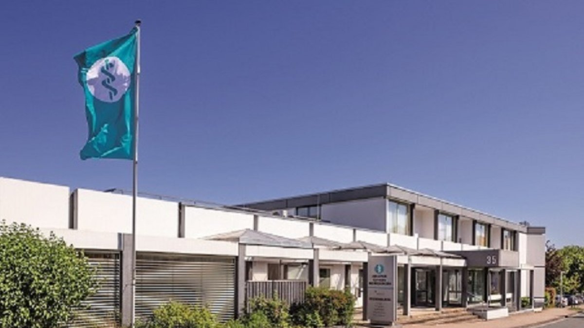 Es ist die vordere Front der Klinik zu sehen. Das Gebäude hat ein Flachdach. Vor dem Eingang der Klinik steht ein Schild, welches die Klinik ausweist. Im Vordergrund steht eine Flagge mit dem Logo von Asklepios.