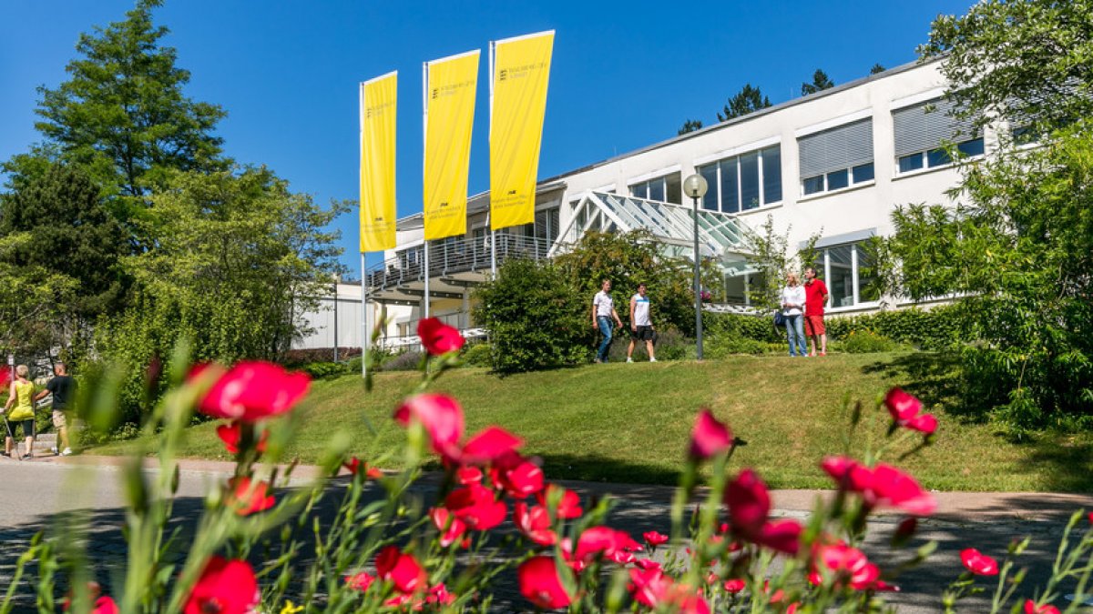 Blick auf den Haupteingang der Klinik Schwabenland in Isny mit drei gelben Klinikfahnen; im Vordergrund blühende Blumen und Bäume – einladendes Ambiente der Rehaklinik