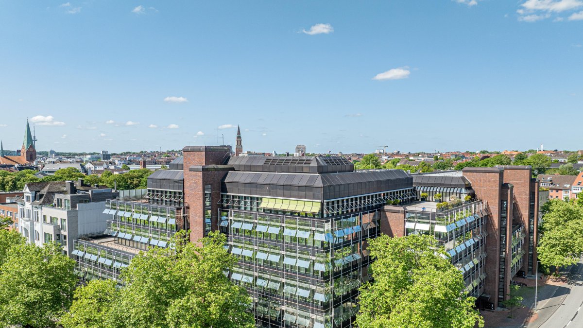Dronenaufnahme vom Gebäude, das vornehmlich aus Glas besteht, mit grünen Bäumen im Vordergrund und der Stadtsilhouette im Hintergrund, bspw. mit dem Rathausturm