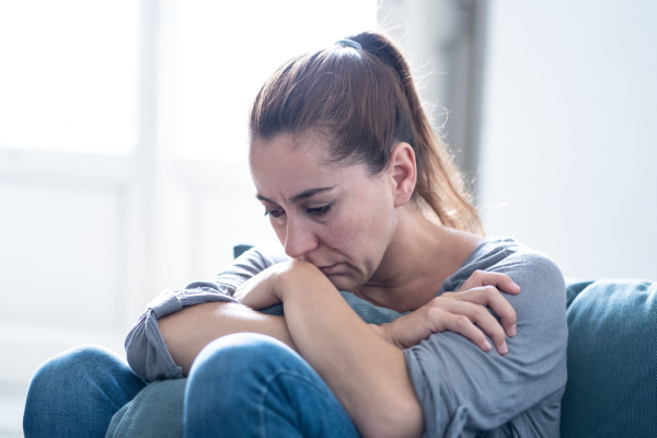 Eine Frau sitzt mit gesenktem Kopf und angewinkelten Beinen auf der Couch, hält ein Kissen umklammert und wirkt niedergeschlagen.