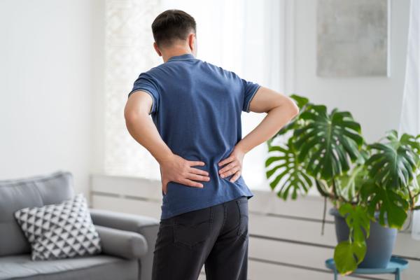 Ein Mann hält sich vor Schmerzen den unteren Rücken und steht im Wohnzimmer – ein typisches Anzeichen für einen Bandscheibenvorfall.