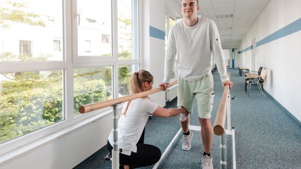 Patient trainiert mit Unterstützung einer Therapeutin an einem Gehbarren in der Rehabilitation.
