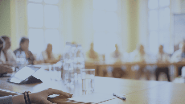 Verschwommenes Bild von Menschen, die um einen runden Konferenztisch sitzen. Hand einer Teilnehmerin im Vordergrund auf der Tischkante.