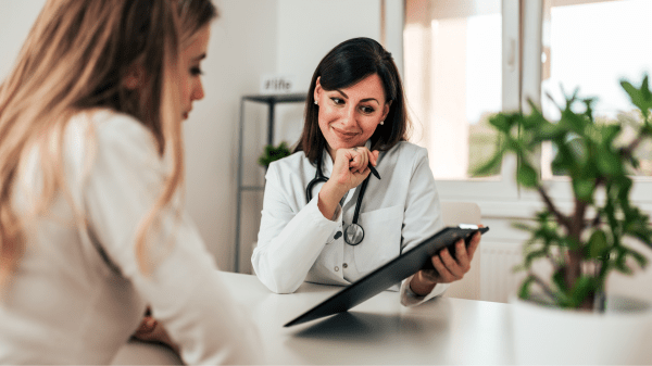 Ärztin mit Klemmbrett im Gespräch mit einer Patientin im hellen Behandlungsraum einer Rehaklinik.