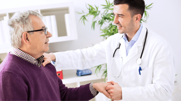 Ein Arzt spricht lächelnd mit einem älteren Patienten und hält dessen Hand. Eine Situation, die Vertrauen und Mitentscheidung symbolisiert.