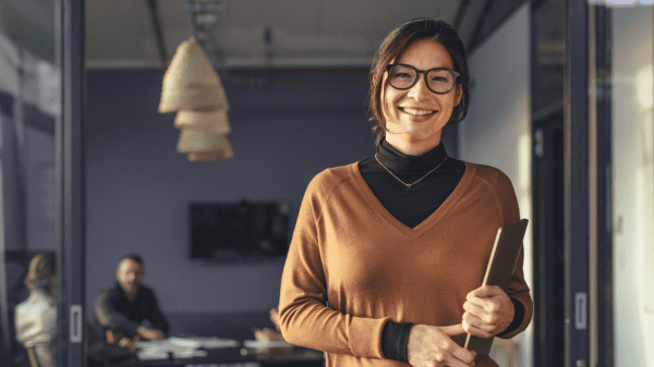 Eine Frau steht in einem modernen Büro und lächelt in Richtung Kamera. Das Bild vermittelt einen gelungenen Wiedereinstieg in den Arbeitsalltag.