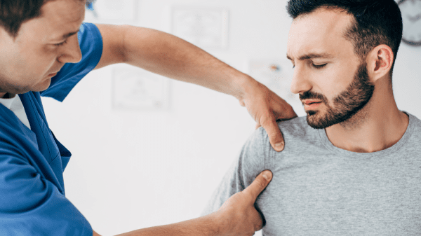 Ein Mann mit schmerzverzerrtem Gesicht wird nach einem Schulterbruch von einem Physiotherapeuten an der Schulter behandelt.