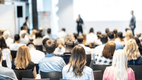 Menschen blicken bei einer Veranstatlung auf eine Bühne, helle Farben, leicht verschwommen
