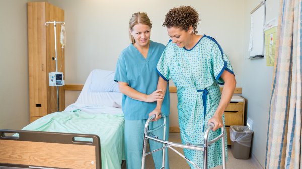 Frau wird nach Operation durch eine Pflegefachkraft beim Laufen unterstützt.