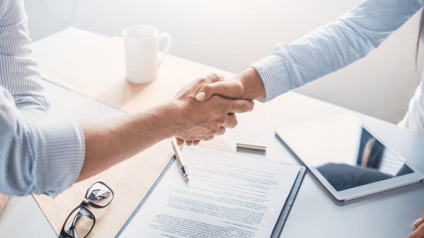 Zwei Personen geben sich über einem Schreibtisch mit Vertragsunterlagen, Brille und Tablet die Hand