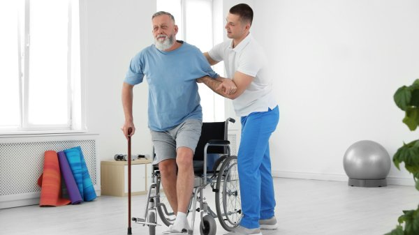 Mann wird Gehhilfe wird durch Physiotherapeut unterstützt aus Rollstuhl aufzustehen im Rahmen einer Anschlussrehabilitation.