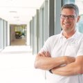 Portrait Dr. Gilbert Rosar lehnt mit verschränkten Armen an Glaswand