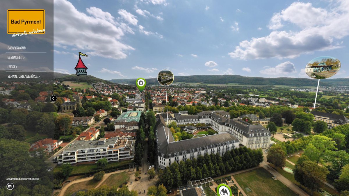 Virtueller Rundgang Bad Pyrmont: Luftaufnahme der Stadt mit Kurpark, historischen Gebäuden und umliegender Landschaft, interaktiven Markierungen zu Sehenswürdigkeiten und Gesundheitseinrichtungen.