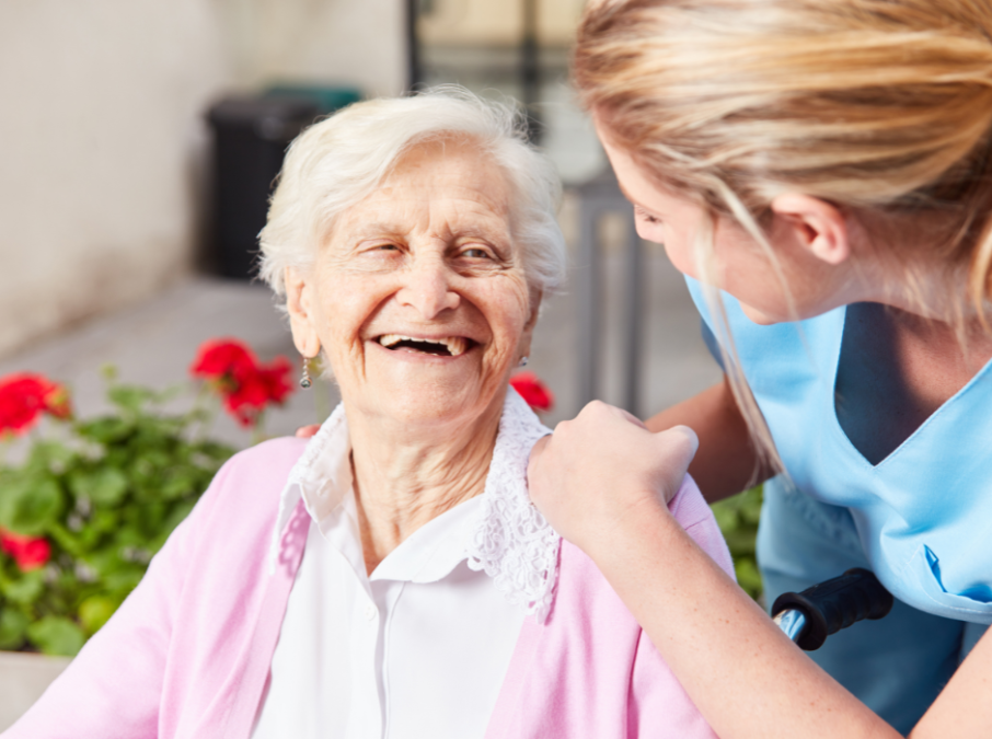 Ältere Patientin lacht Pflegerin an, die sich über den Stuhl herunter beugt