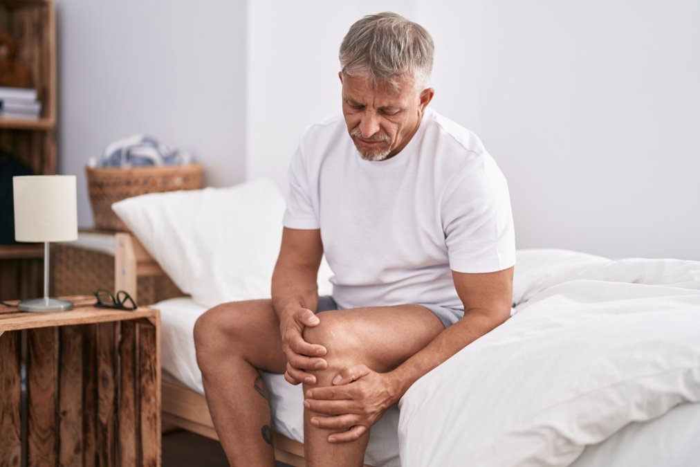 Ein Mann sitzt auf der Bettkante und hält sich mit schmerzverzerrtem Gesicht das Knie, ein typisches Symptom von Arthrose.