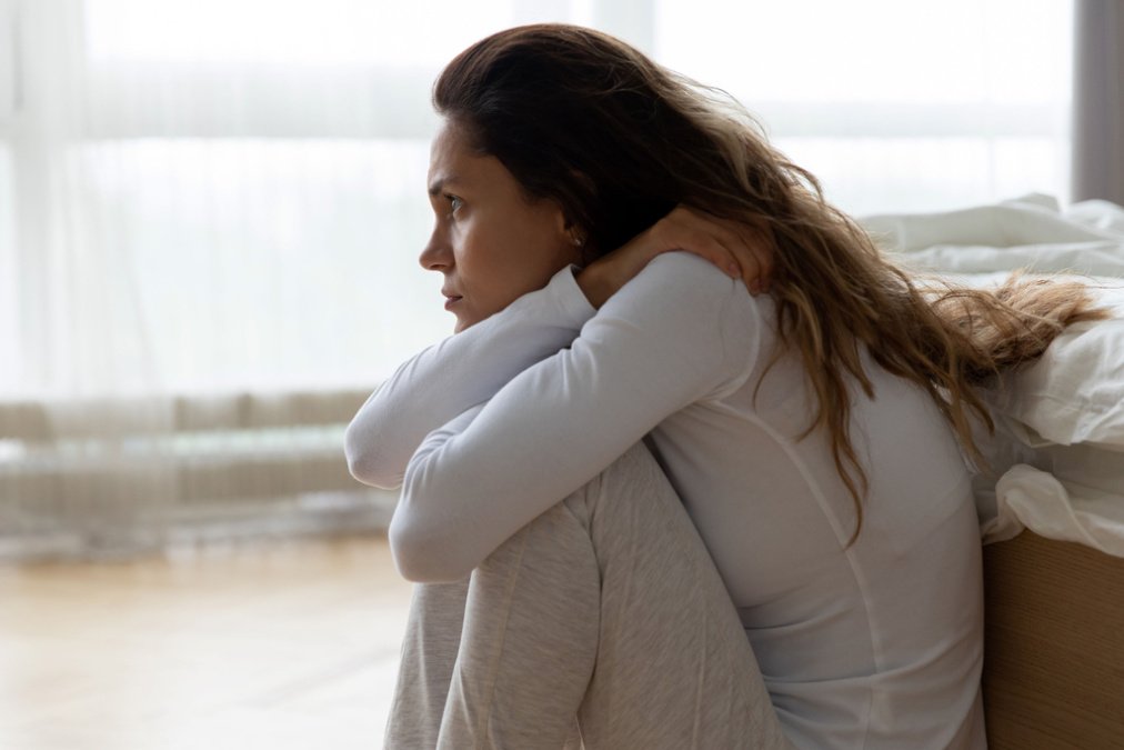 Eine verängstigte Frau sitzt in der Hocke auf dem Boden vor einem Bett, sichtlich belastet von einer Angststörung.