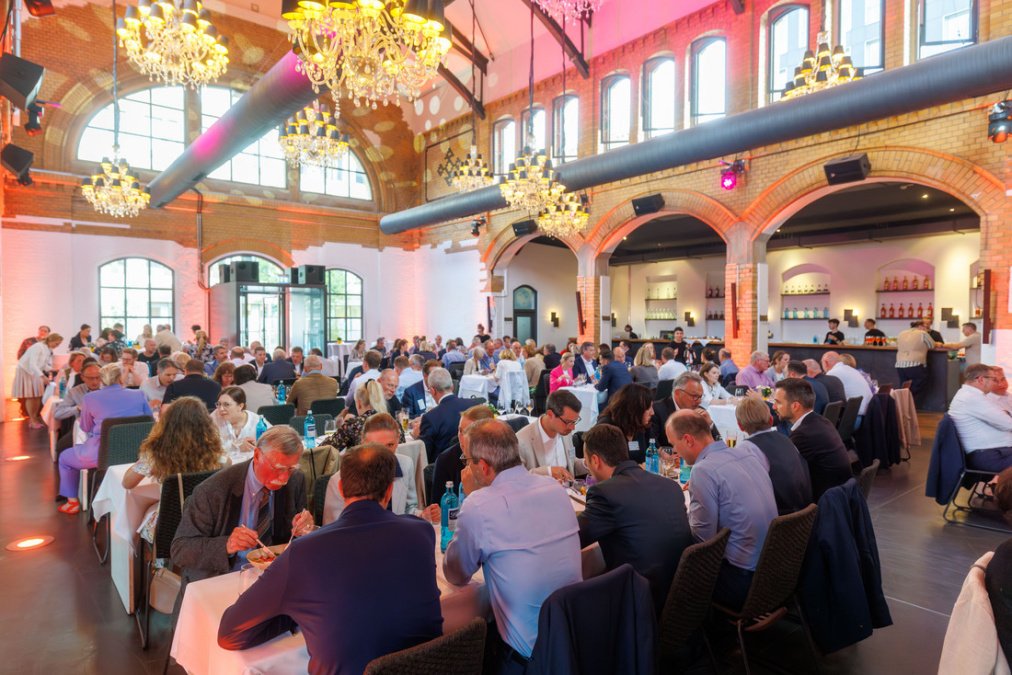 Wartehalle ausgeleuchtet in bunten Farben. Gäste sitzen in langen Tischen beim Essen.