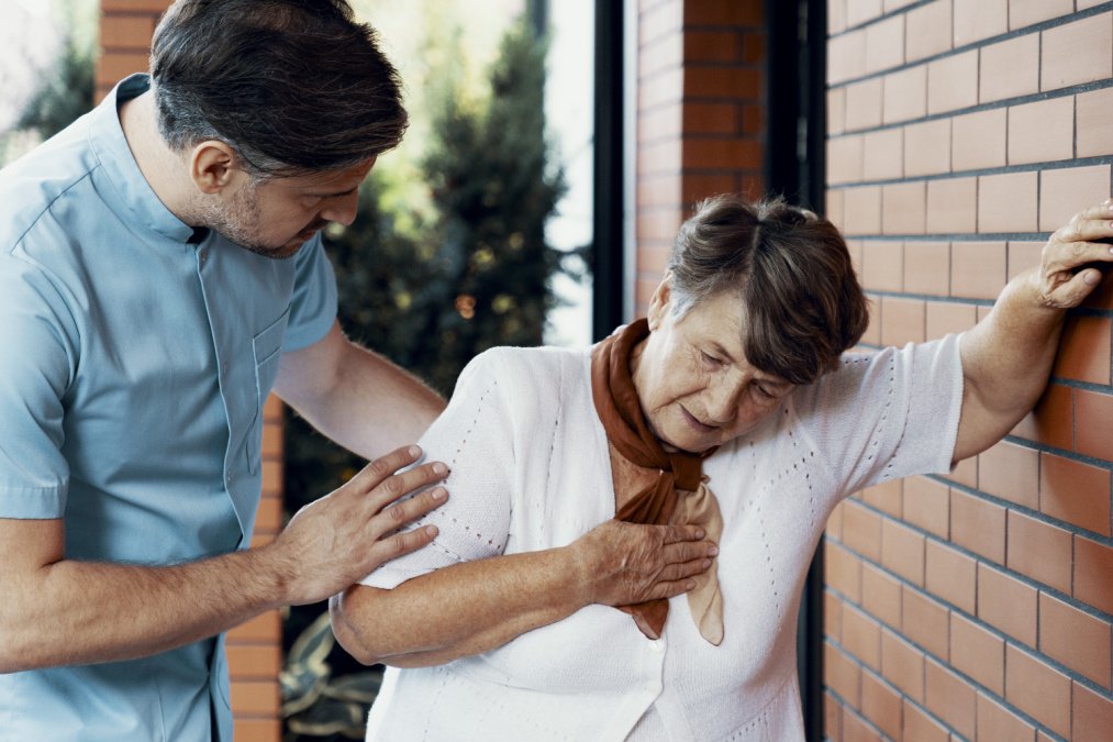 Eine Frau lehnt sich gegen die Wand, eine Hand auf ihrer Brust, während sie von einem Mann neben ihr unterstützt wird.