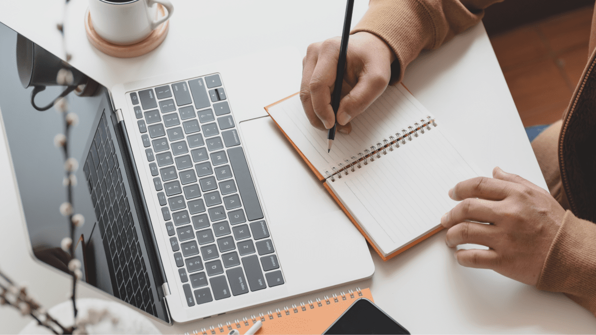 Draufsicht auf eine Person, die mit einem Stift in ein Notizbuch schreibt. Daneben stehen ein geöffneter Laptop und eine Kaffeetasse.