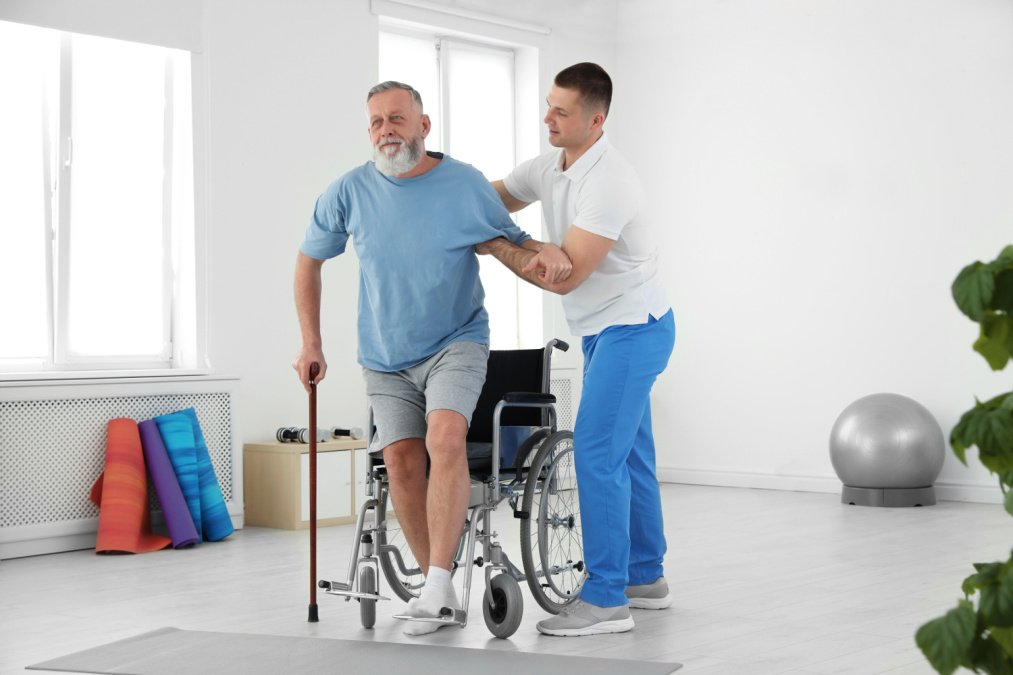 Mann wird Gehhilfe wird durch Physiotherapeut unterstützt aus Rollstuhl aufzustehen im Rahmen einer Anschlussrehabilitation.